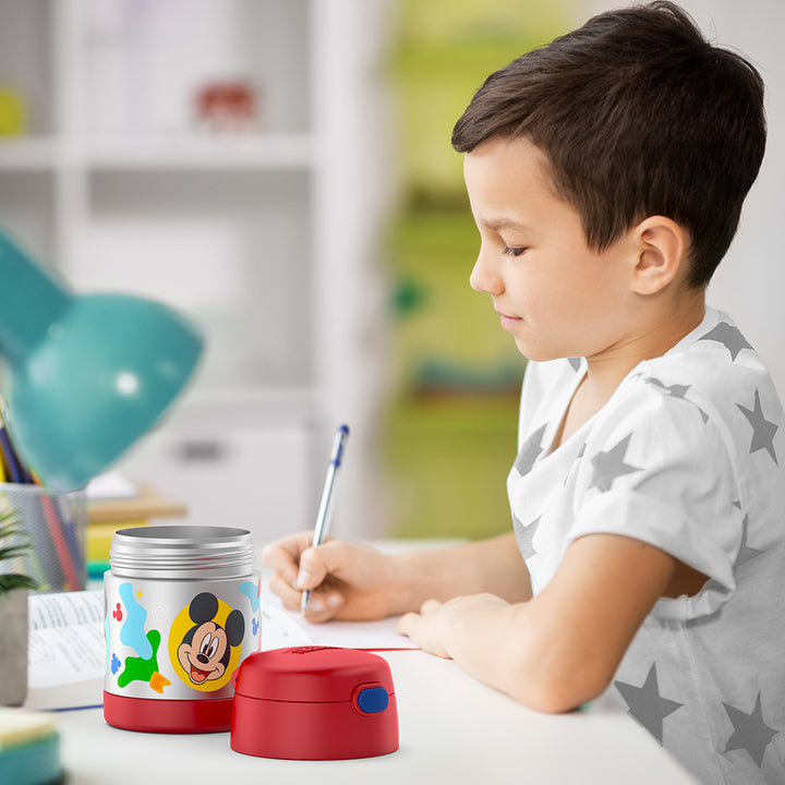 Boy sitting at table doing homework with an open 10 ounce Mickey Mouse Funtainer food jar open next to him.