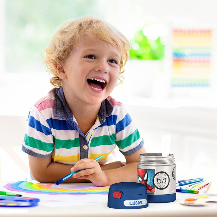 Boy smiling, sitting at table painting with an open 10 ounce Spider-Man Funtainer food jar open in front of him. 
