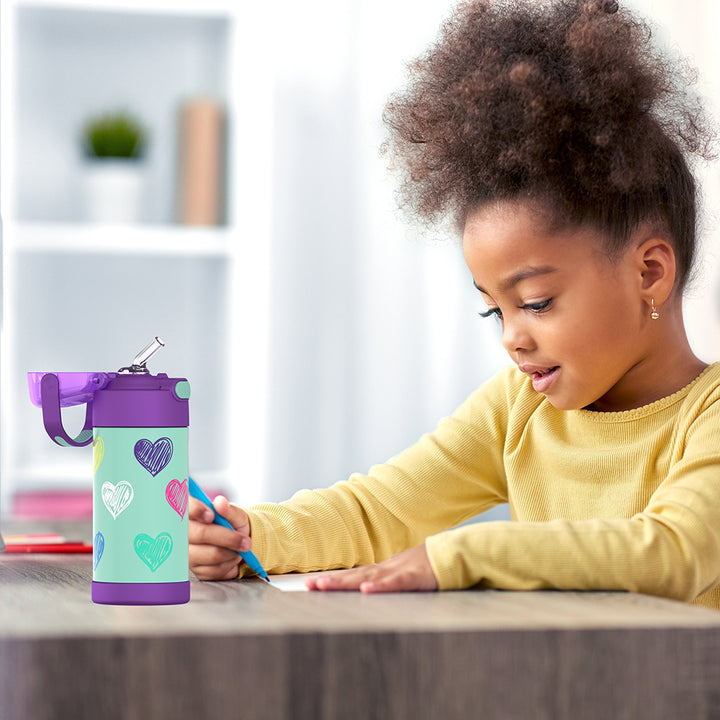 Girl sitting at a table doing homework, with a 12 ounce colorful sketched Hearts Pattern Funtainer water bottle opened next to her.