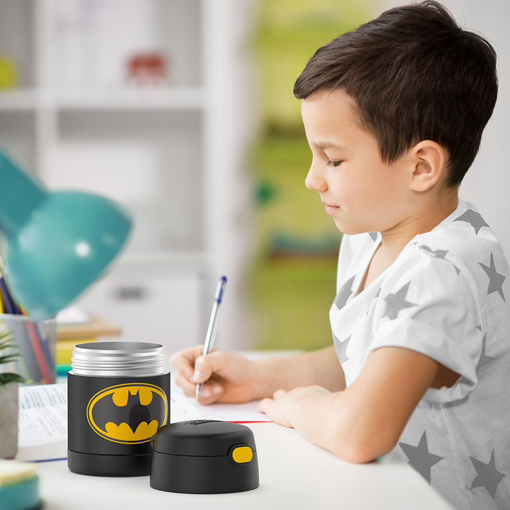 Boy sitting at table doing homework with an open 10 ounce Batman Funtainer food jar open next to him.