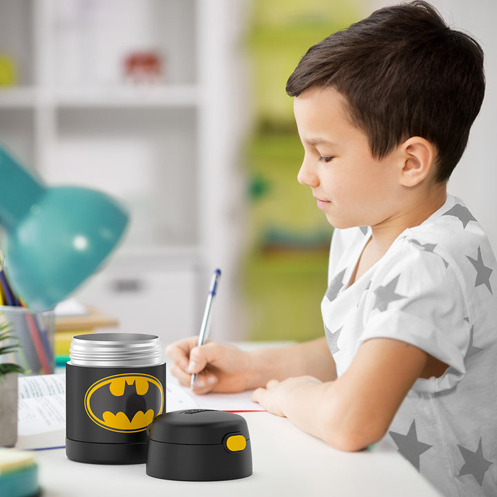 Boy sitting at table doing homework with an open 10 ounce Batman Funtainer food jar open next to him.