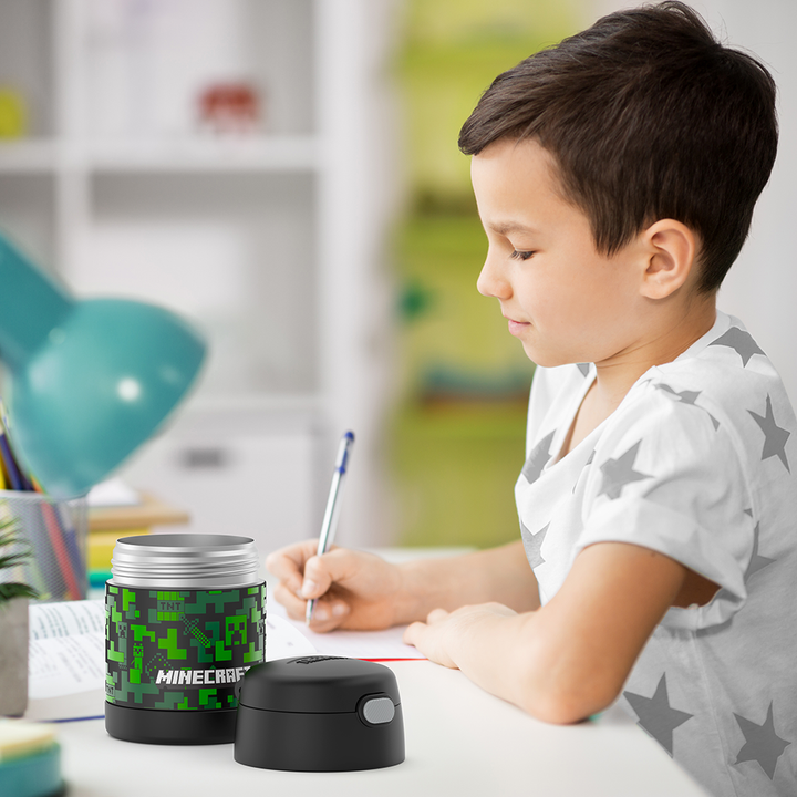Boy sitting at table doing homework with an open 10 ounce Minecraft Funtainer food jar open next to him.