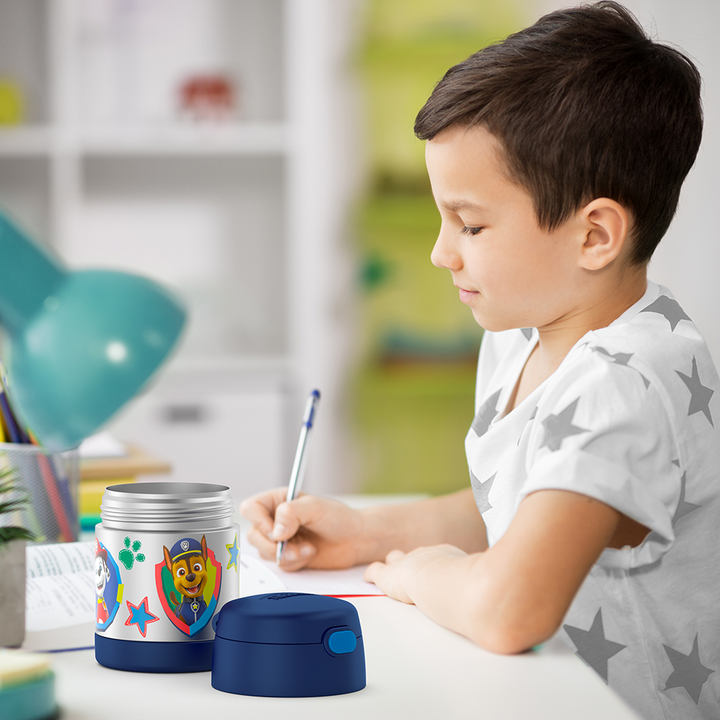 Boy sitting at table doing homework with an open 10 ounce Paw Patrol Funtainer food jar open next to him.