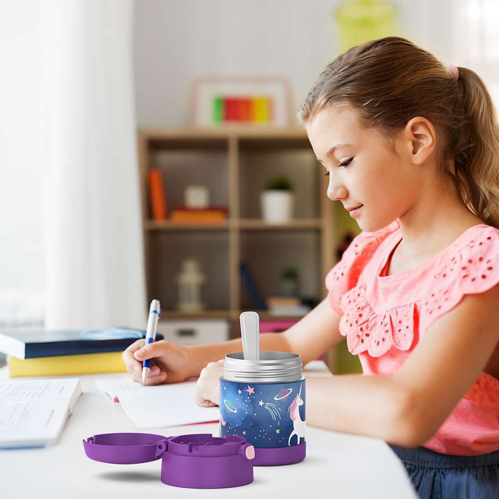 Girl sitting at table doing homework with an open 10 ounce Funtainer unicorn in space food jar open next to her.